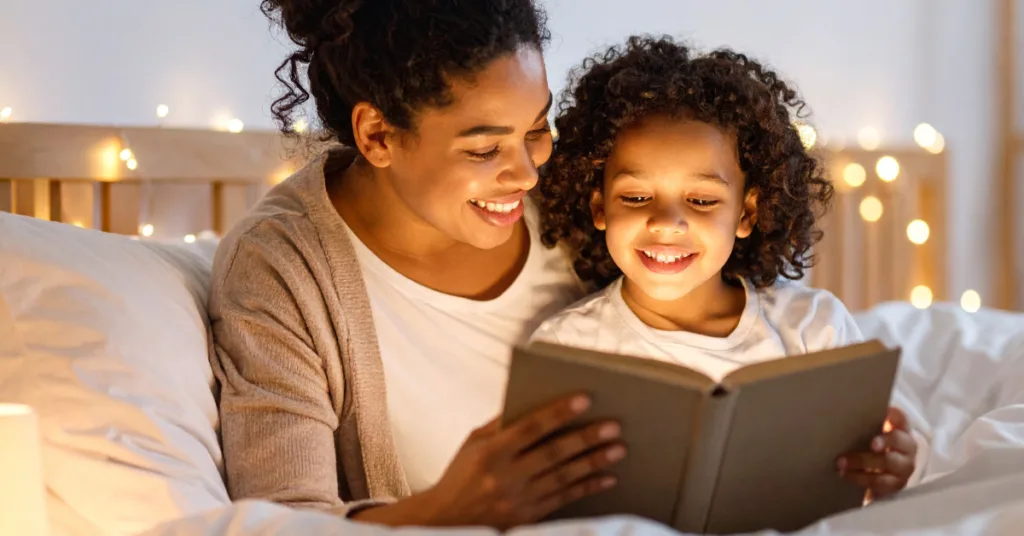 Mom and daughter reading a book and smiling in bed with fairy lights